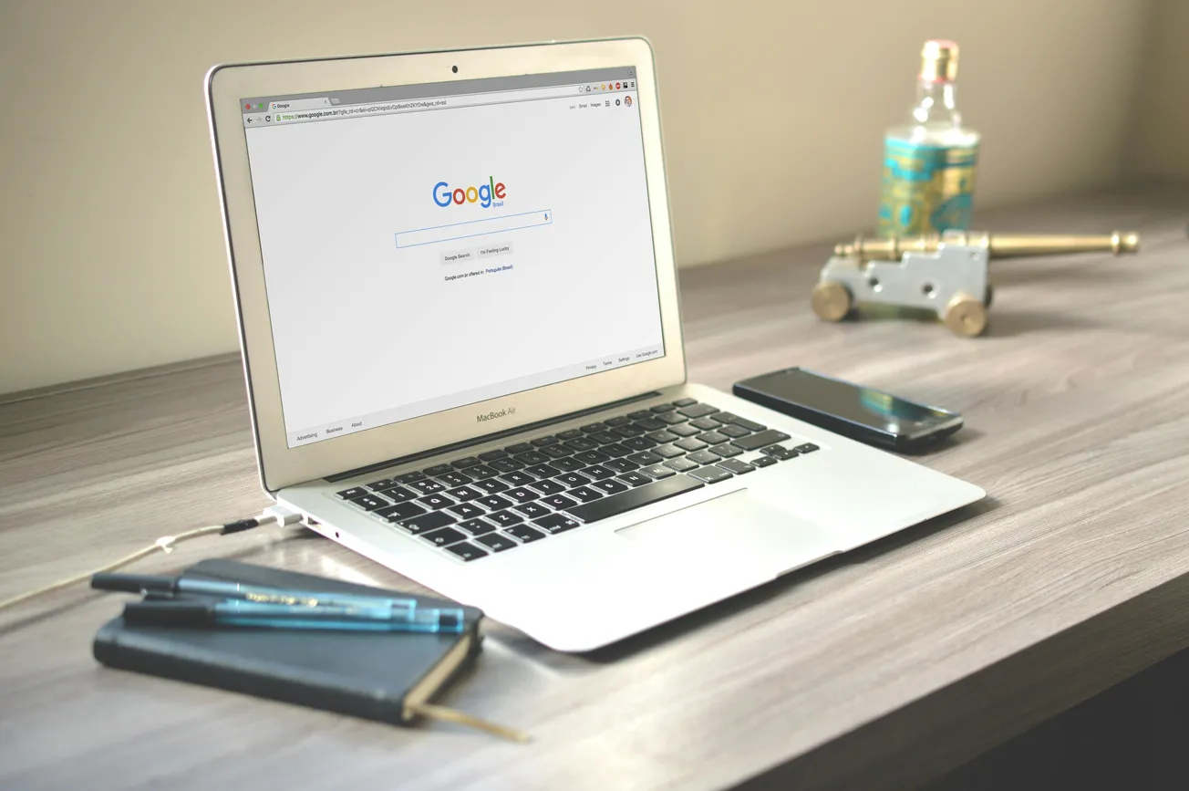 A laptop displaying Google's homepage sits on a wooden desk. Beside it are a notebook, pen, smartphone, and decorative items.