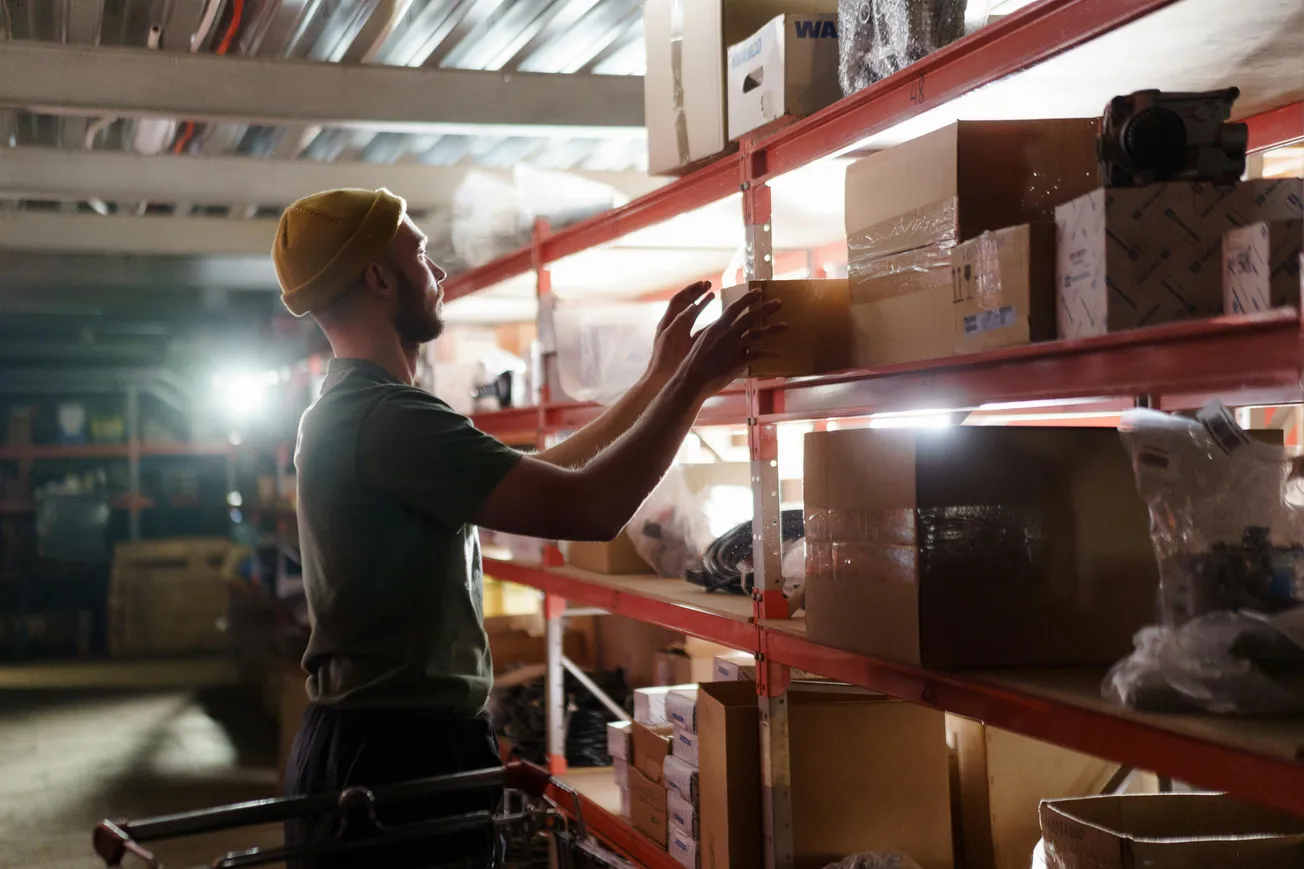 A man wearing a yellow beanie and green shirt reaches for boxes on red metal shelves in a dimly lit warehouse, creating a focused and industrious mood.