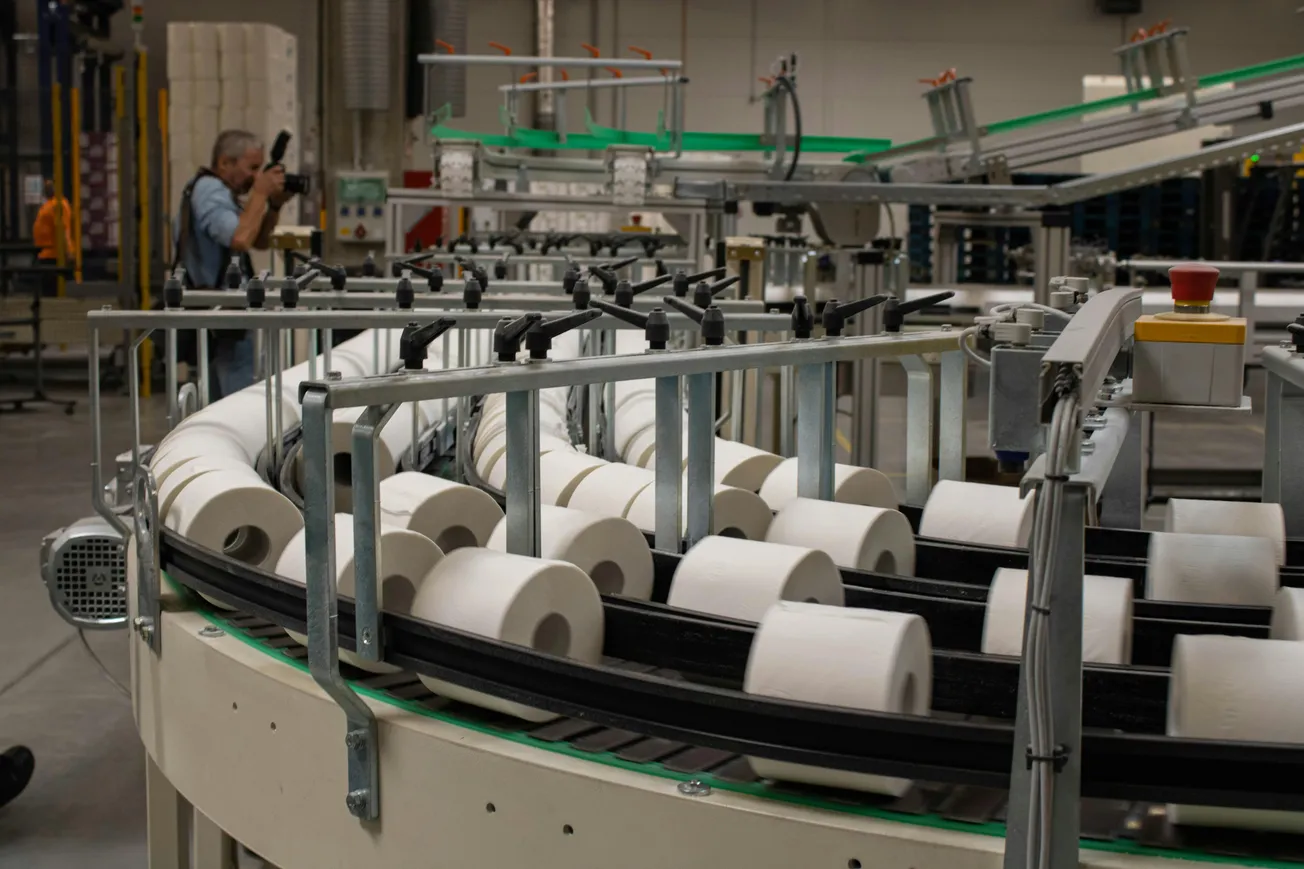 A factory setting featuring a conveyor belt with rolls of toilet paper. A person on the left captures the scene with a camera.