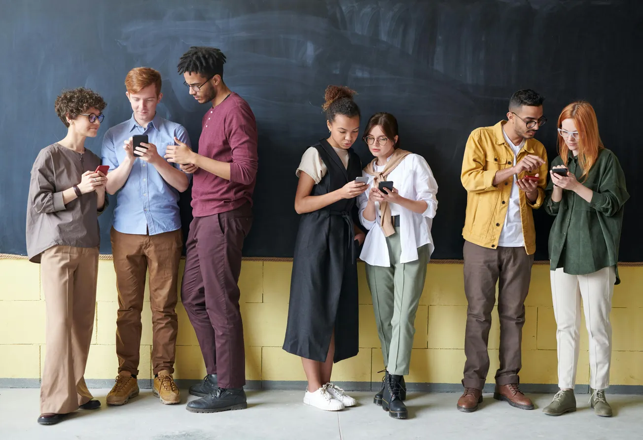 A group of seven people standing in a line against a blackboard, focused on their smartphones, conveying a relaxed and engaged atmosphere.