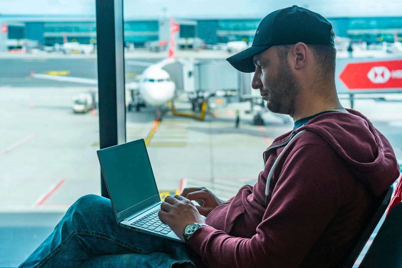 A man in a cap and jacket sits by an airport window, focused on his laptop. A plane is visible outside, conveying a busy, travel-focused atmosphere.