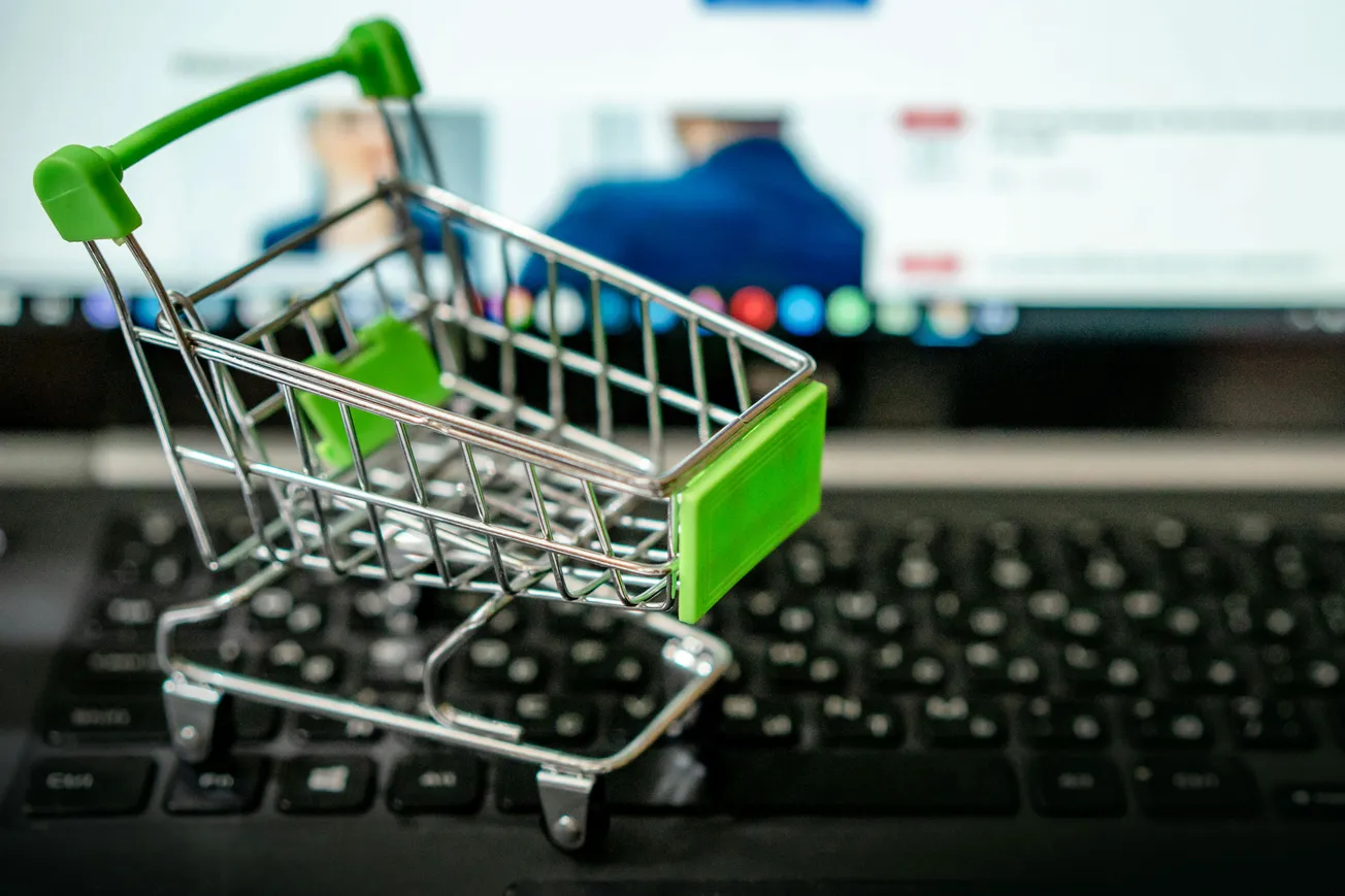 A small shopping cart with green accents sits atop a keyboard, symbolizing online shopping. A blurred screen is visible in the background.