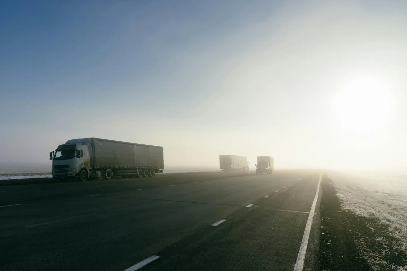 Foggy highway scene with three trucks driving towards a distant horizon. The sun is low and hazy, casting a soft glow.