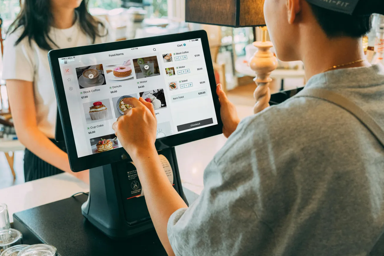 A person in a cap uses a touchscreen at a coffee shop, selecting items like pastries and drinks. The atmosphere is casual and modern.