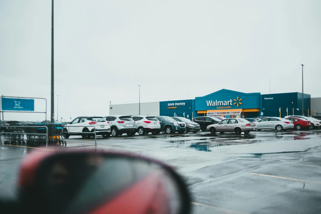 Parking lot of a Walmart store on a rainy day, with wet pavement and rows of parked cars. A large Walmart building is visible in the background.