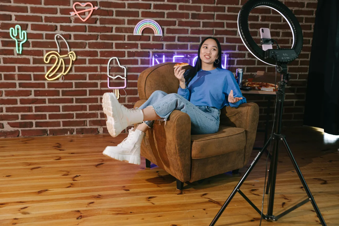 A woman sits in a cozy chair, holding pizza, facing a ring light and phone. She is in a room with neon decor against a brick wall, conveying a casual, creative vibe.