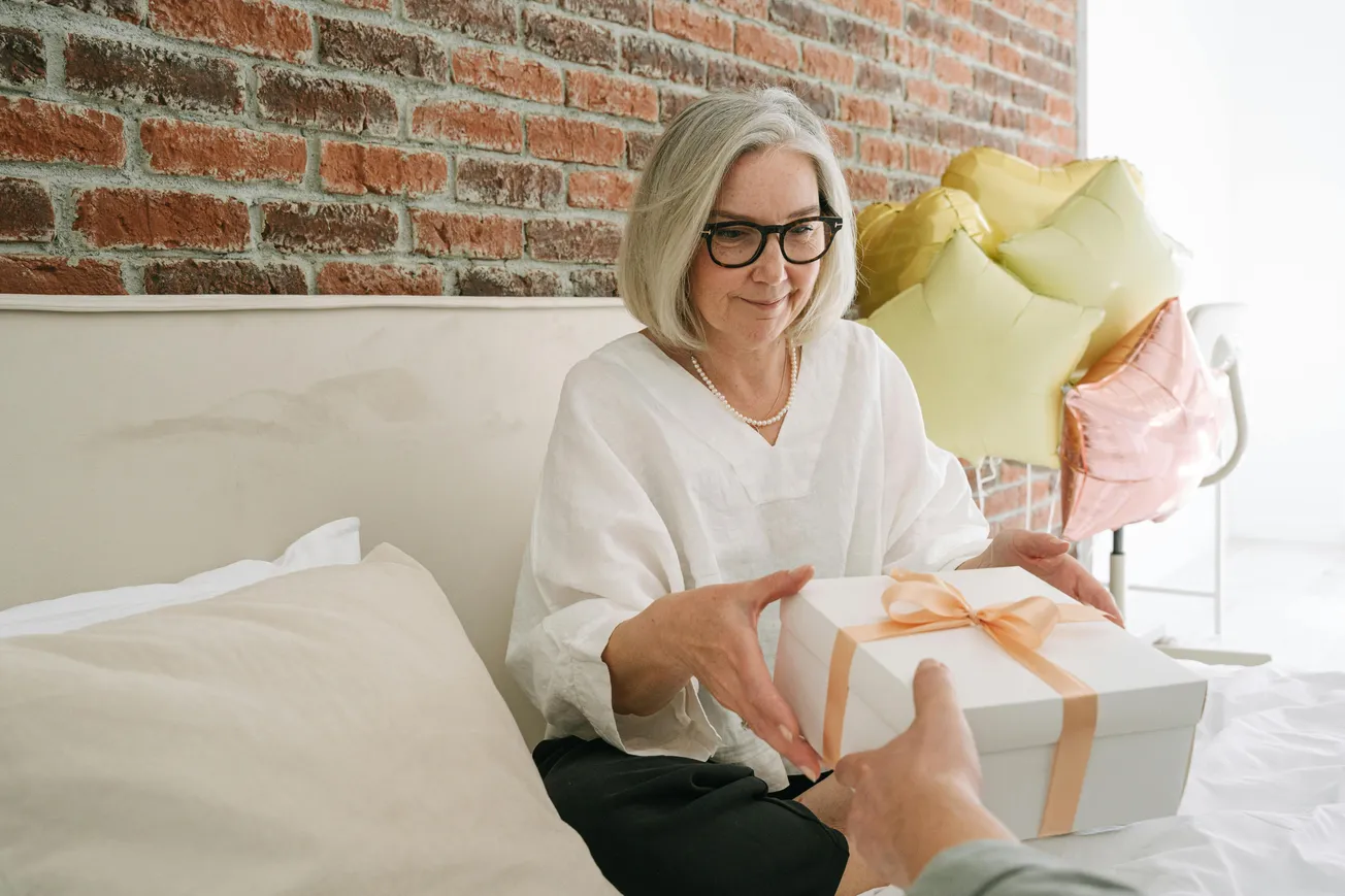 Elderly woman with glasses, wearing a white blouse, smiles while receiving a gift box with a peach ribbon. Heart-shaped balloons decorate the brick wall.