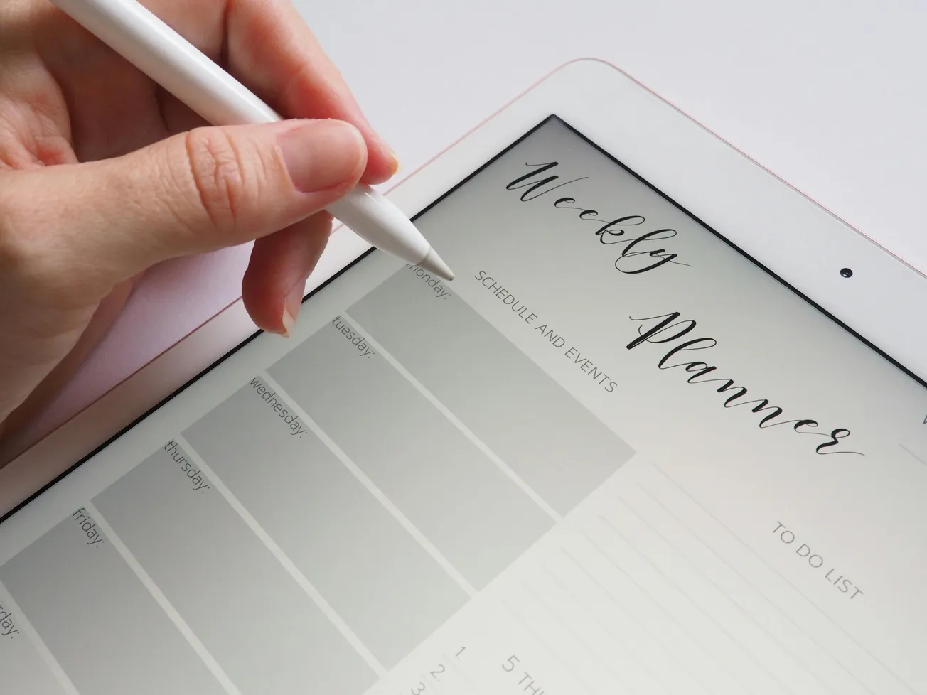 A hand holds a stylus over a digital tablet displaying a weekly planner. The planner has neatly arranged days and sections for scheduling and tasks.