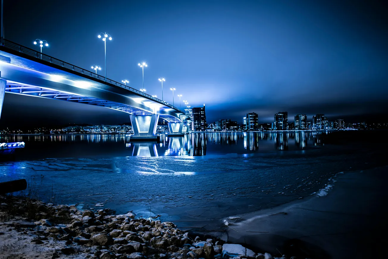 Illuminated bridge reflecting on a calm river at night, with city skyline in the background. The scene conveys a serene, futuristic atmosphere.