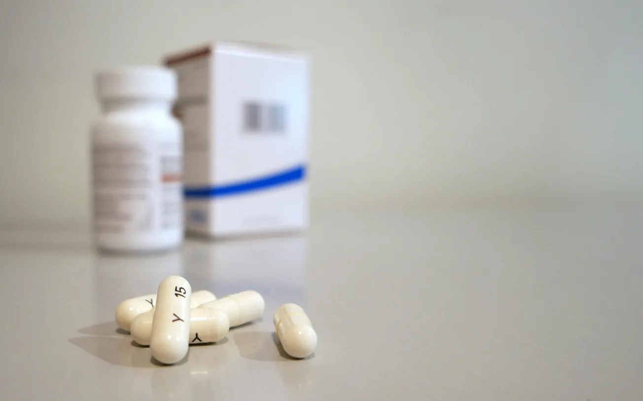 White capsules labeled "Y 15" are scattered on a light surface. A blurred prescription bottle and box are in the background, conveying a medical theme.