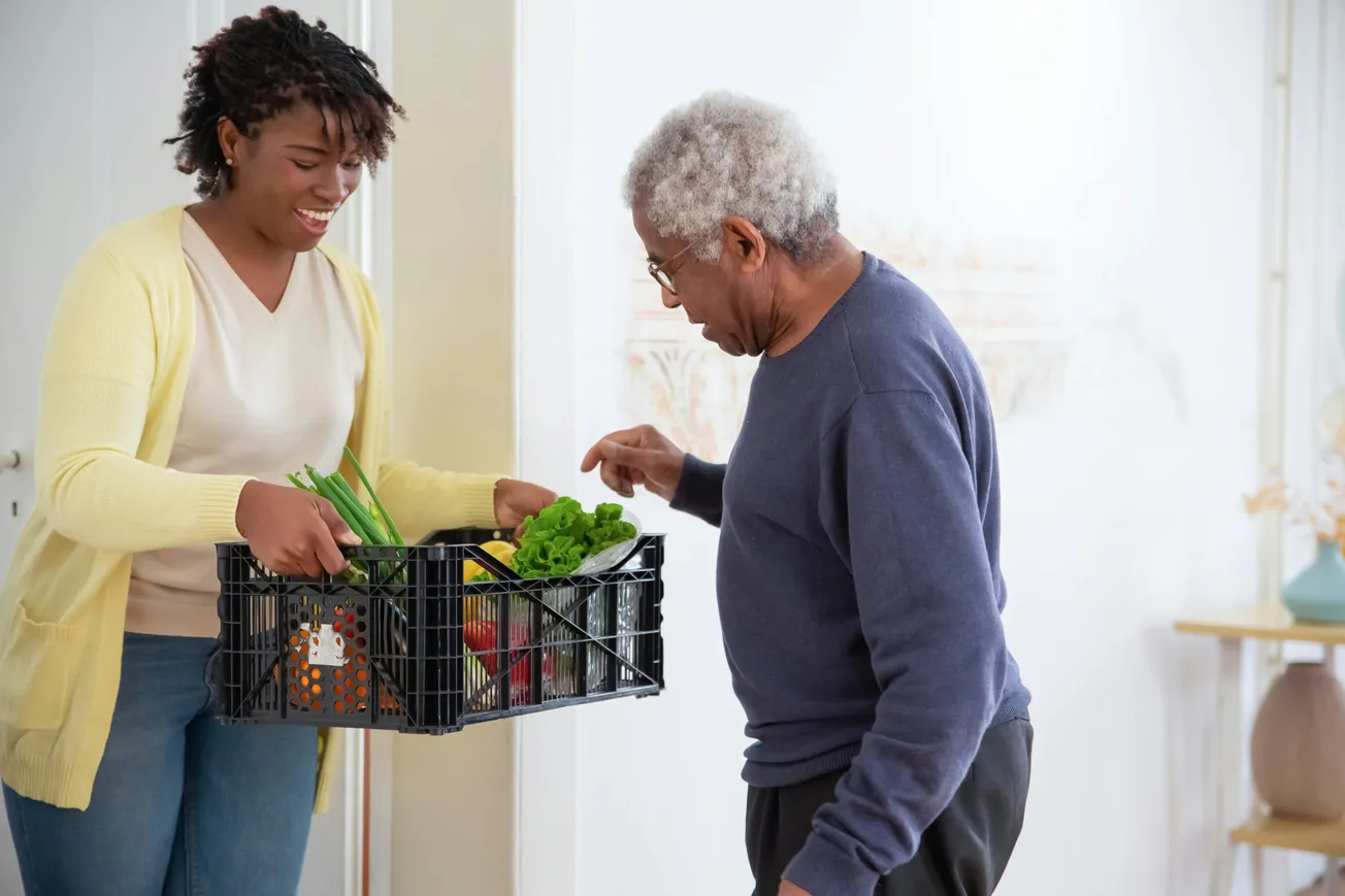A smiling woman in a yellow cardigan hands a black crate of fresh vegetables to an elderly man in a blue sweater.
