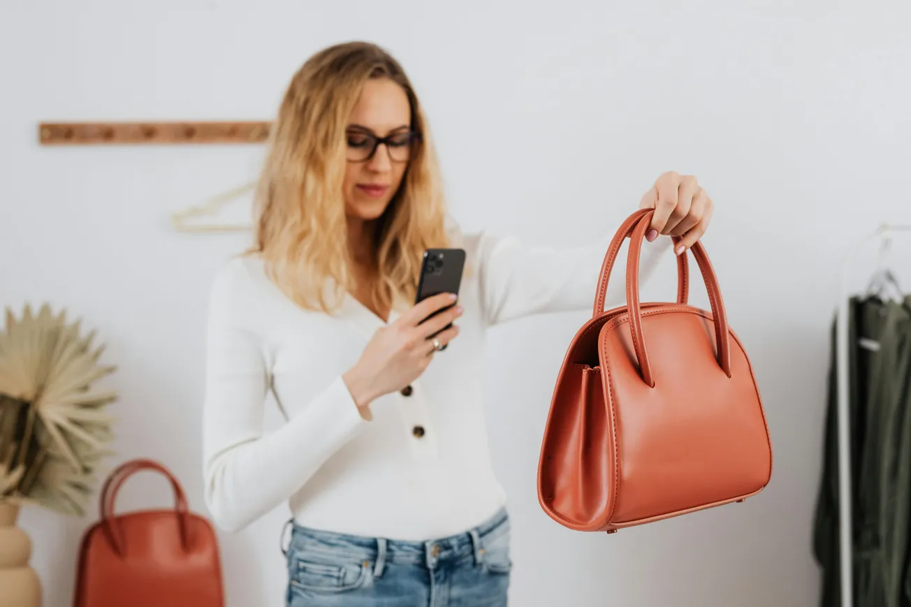A woman in a white top holds a brown handbag in one hand and takes a photo with a smartphone in the other.
