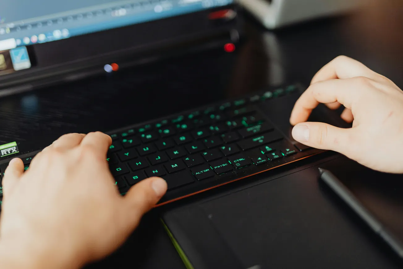 Hands typing on a keyboard with green backlighting. A laptop screen is visible, suggesting a focused, tech-driven work environment.