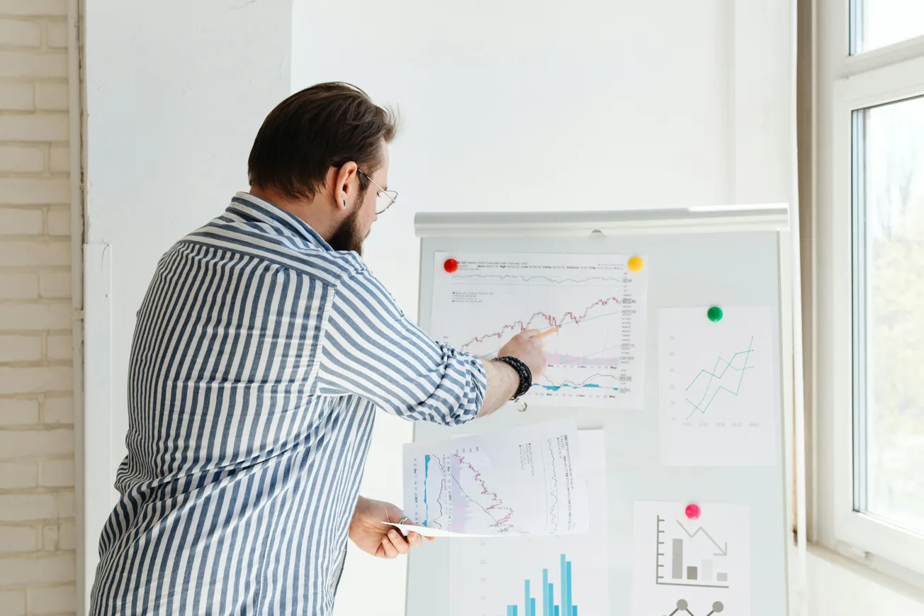 A man in a striped shirt points at graphs on a whiteboard in a bright room. The scene conveys focus and analysis in a business or academic setting.