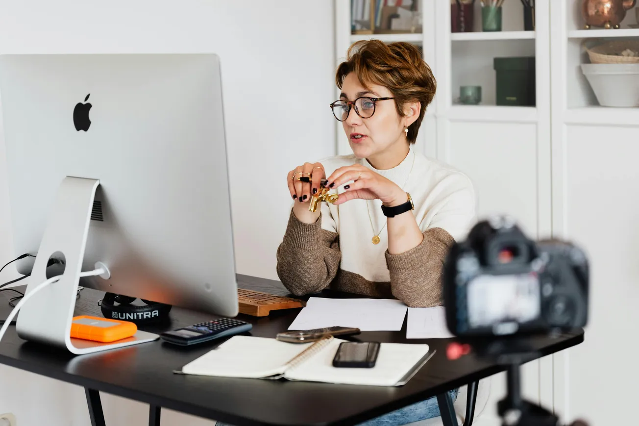 Woman in glasses sits at a desk with a computer and camera, appearing focused during an online meeting. Documents and a smartphone are on the desk.