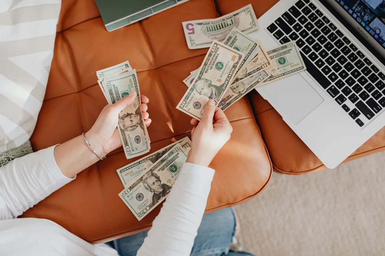 A person counts U.S. dollar bills while sitting on a brown quilted surface, with an open laptop nearby.