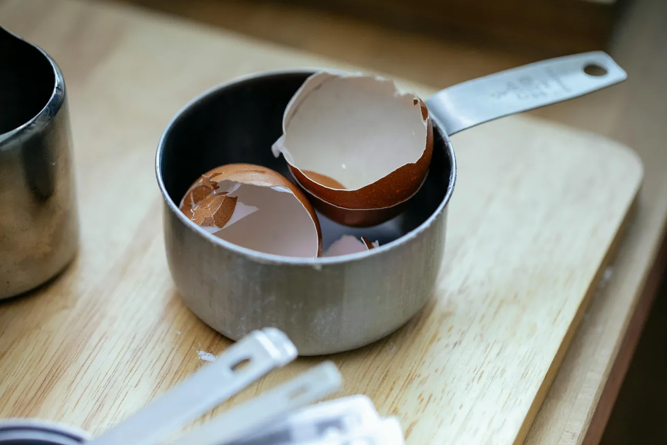 A metal measuring cup on a wooden surface holds broken brown eggshells. The scene conveys a sense of cooking or baking preparation.