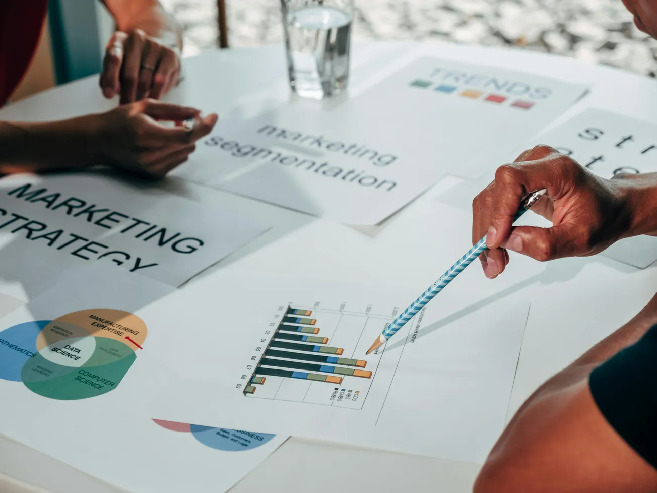 Close-up of hands at a table with charts and documents on marketing strategy and segmentation. A pencil points at a bar graph, conveying analysis.