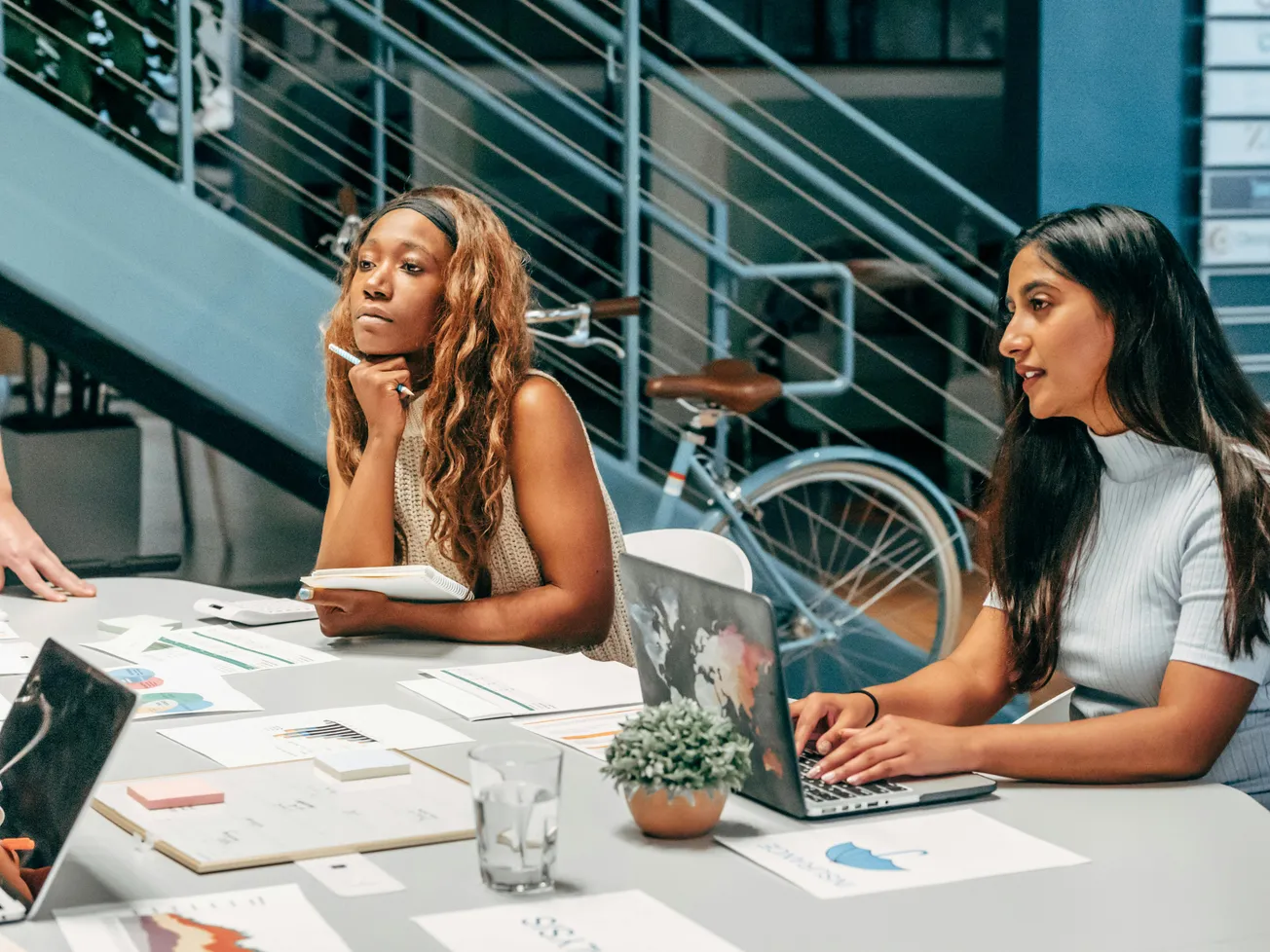 Two women sit at a conference table engaged in discussion. One writes in a notebook, while the other types on a laptop. Papers and a plant are on the table.