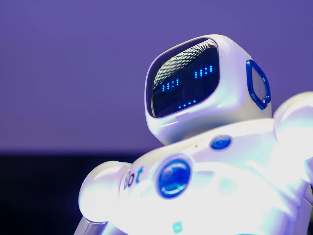 A white robot with a glossy finish is shown against a purple backdrop. Its screen face displays blue lights resembling eyes. The mood is futuristic and friendly.