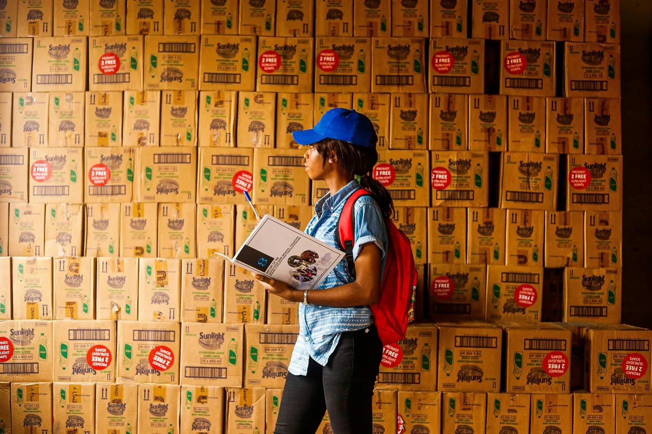 A woman in a blue cap and red backpack holds a clipboard, standing in front of stacked cardboard boxes labeled "Sunlight." The scene is industrious.