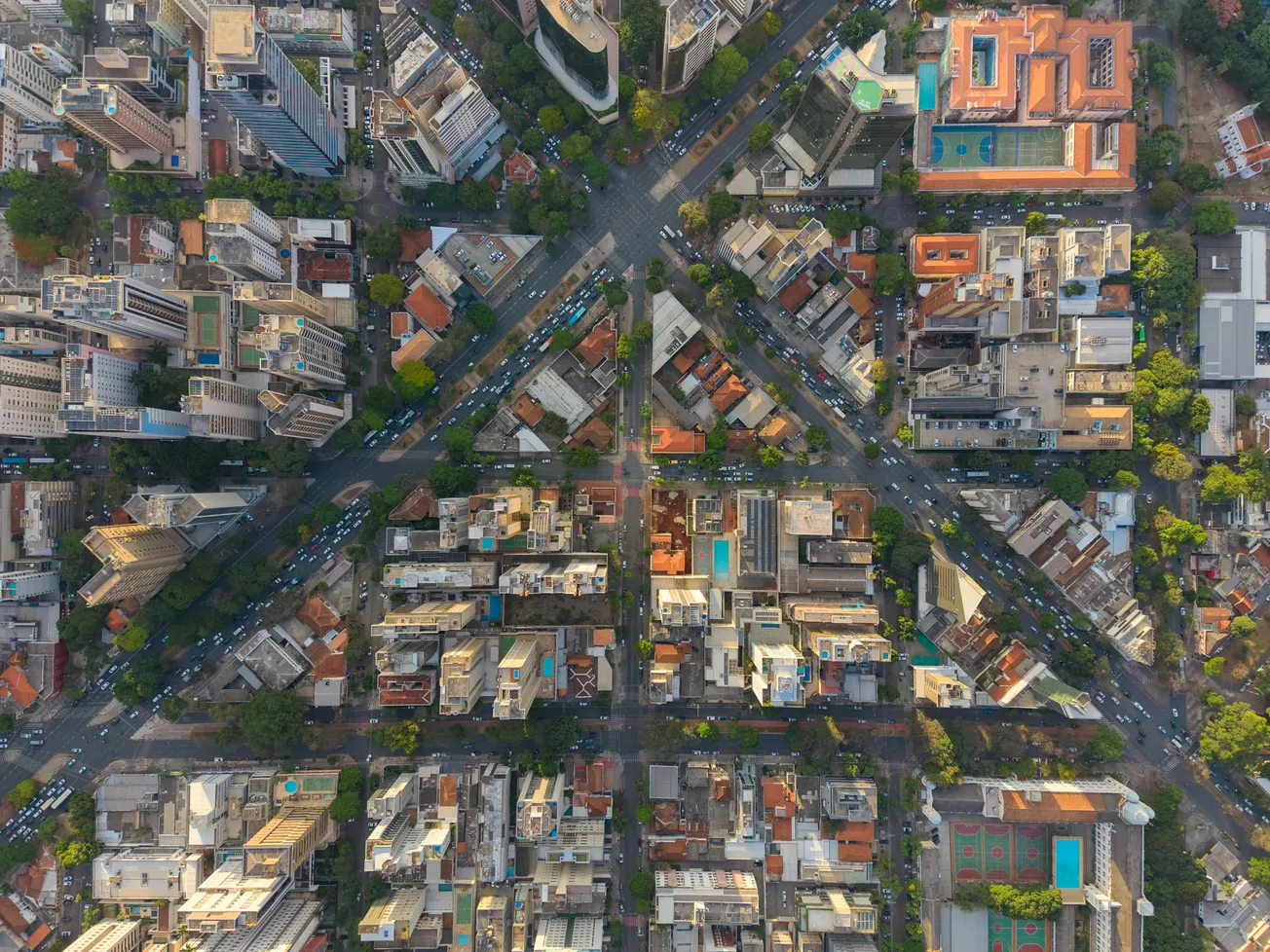 Aerial view of a city intersection forming an 'X', lined with buildings, trees, and streets. Rooftop pools and structures create a dense urban pattern.