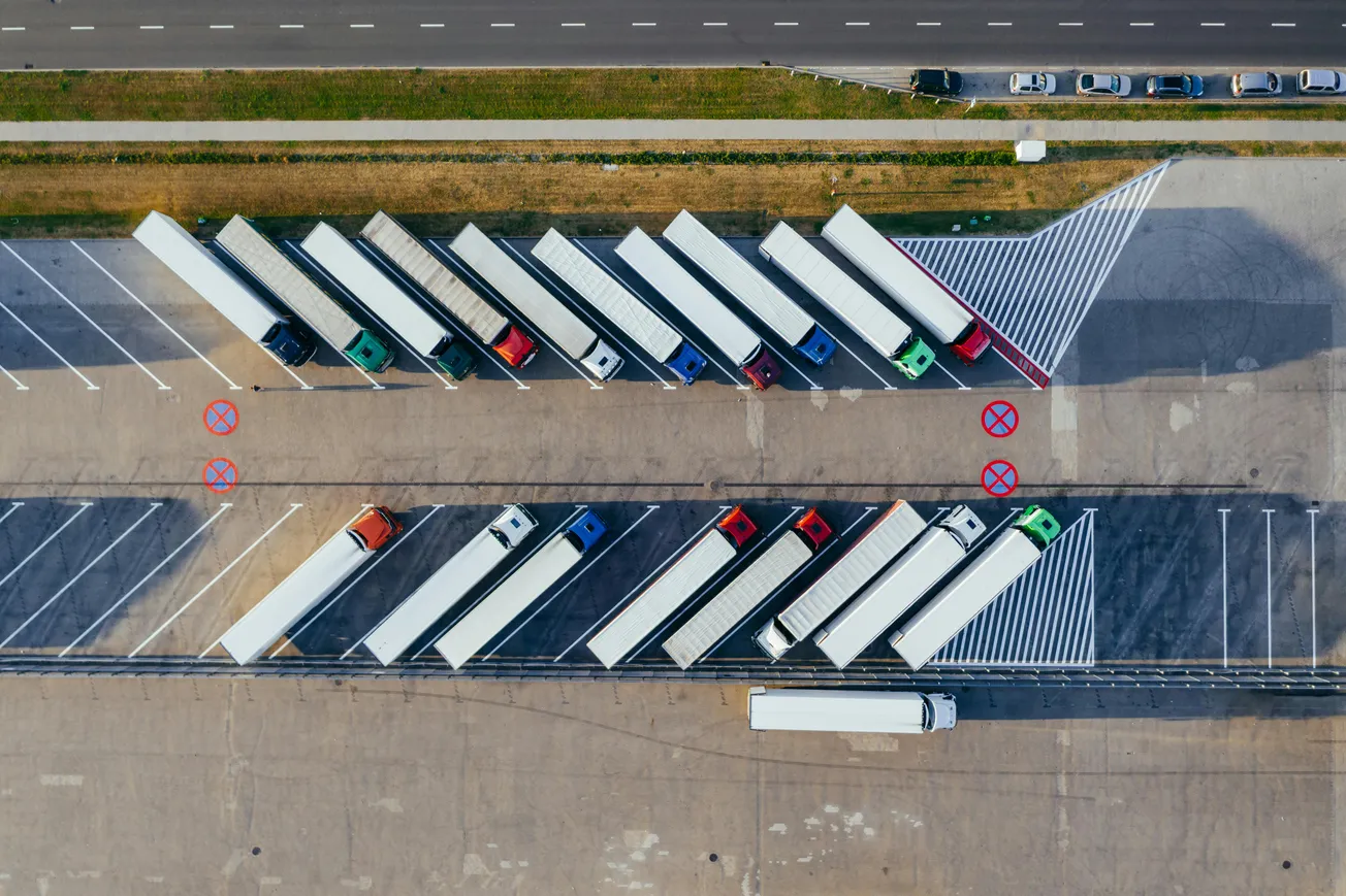 Aerial view of a parking lot with semi-trucks parked diagonally in designated spaces. Red and green truck cabs align neatly. Cars are on the road nearby.