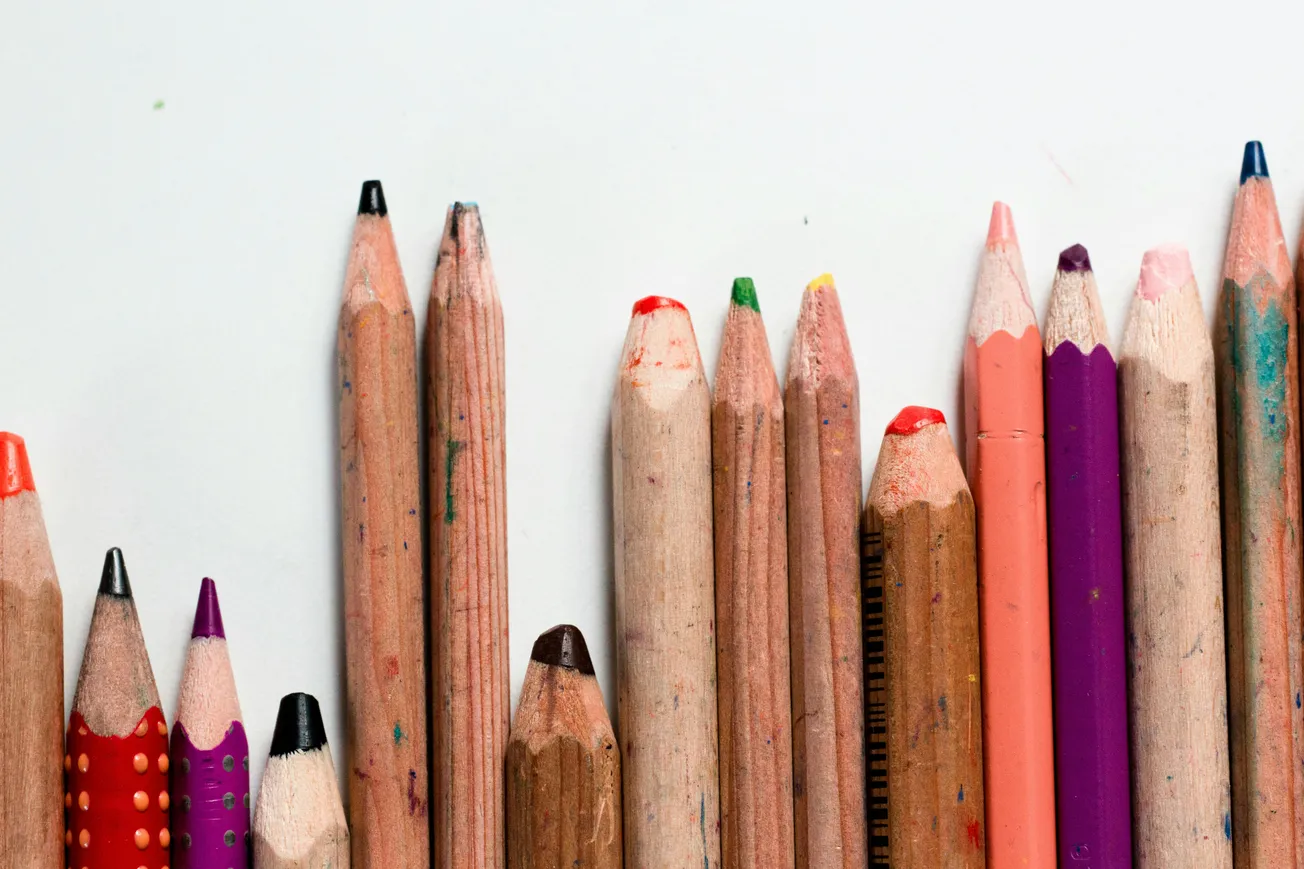 A row of colored pencils, varying in size and color, against a white background. Some have chewed tips and vibrant shells, suggesting creativity and use.