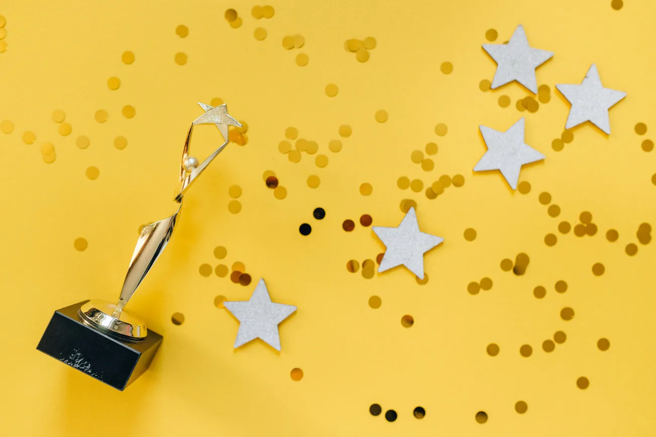 Golden trophy with a star on a black base, surrounded by scattered small gold confetti and five white star cutouts on a bright yellow background.