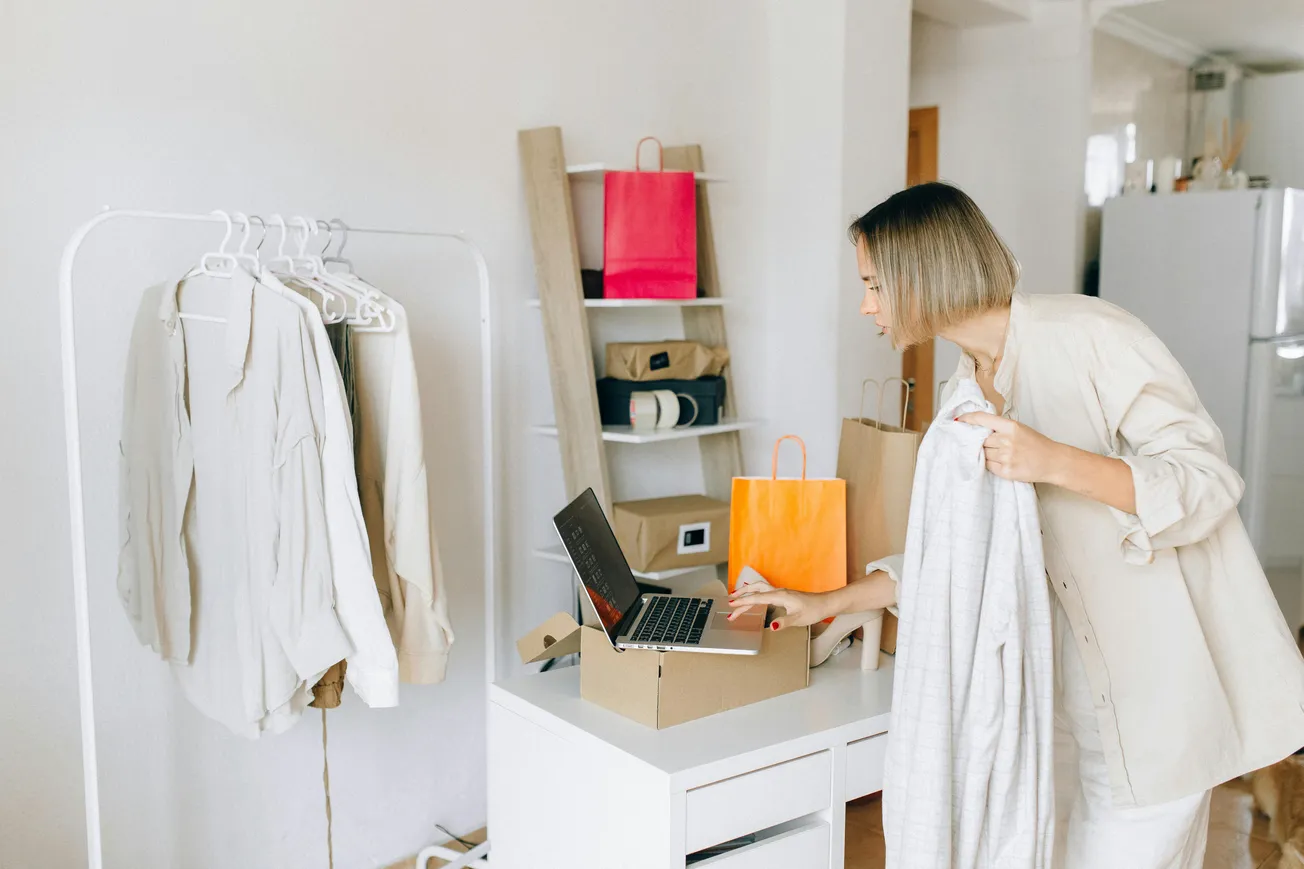 A woman in a light outfit examines a laptop on a desk, holding a garment. Nearby, clothes hang on a rack, and colorful shopping bags are on a shelf.
