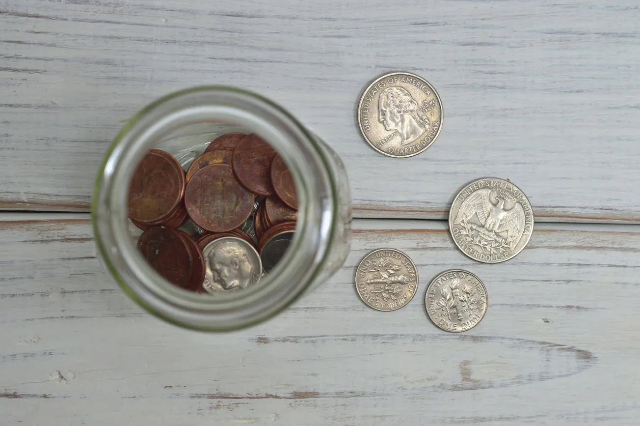 Overhead view of a glass jar filled with pennies on a wooden surface. Nearby are five scattered silver coins, evoking a casual, saving-themed tone.