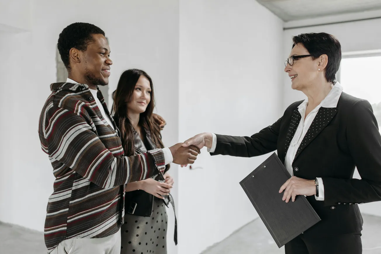 A man and woman stand in a bright, empty room, smiling as the man shakes hands with a professional woman in a suit holding a clipboard, conveying a tone of agreement.