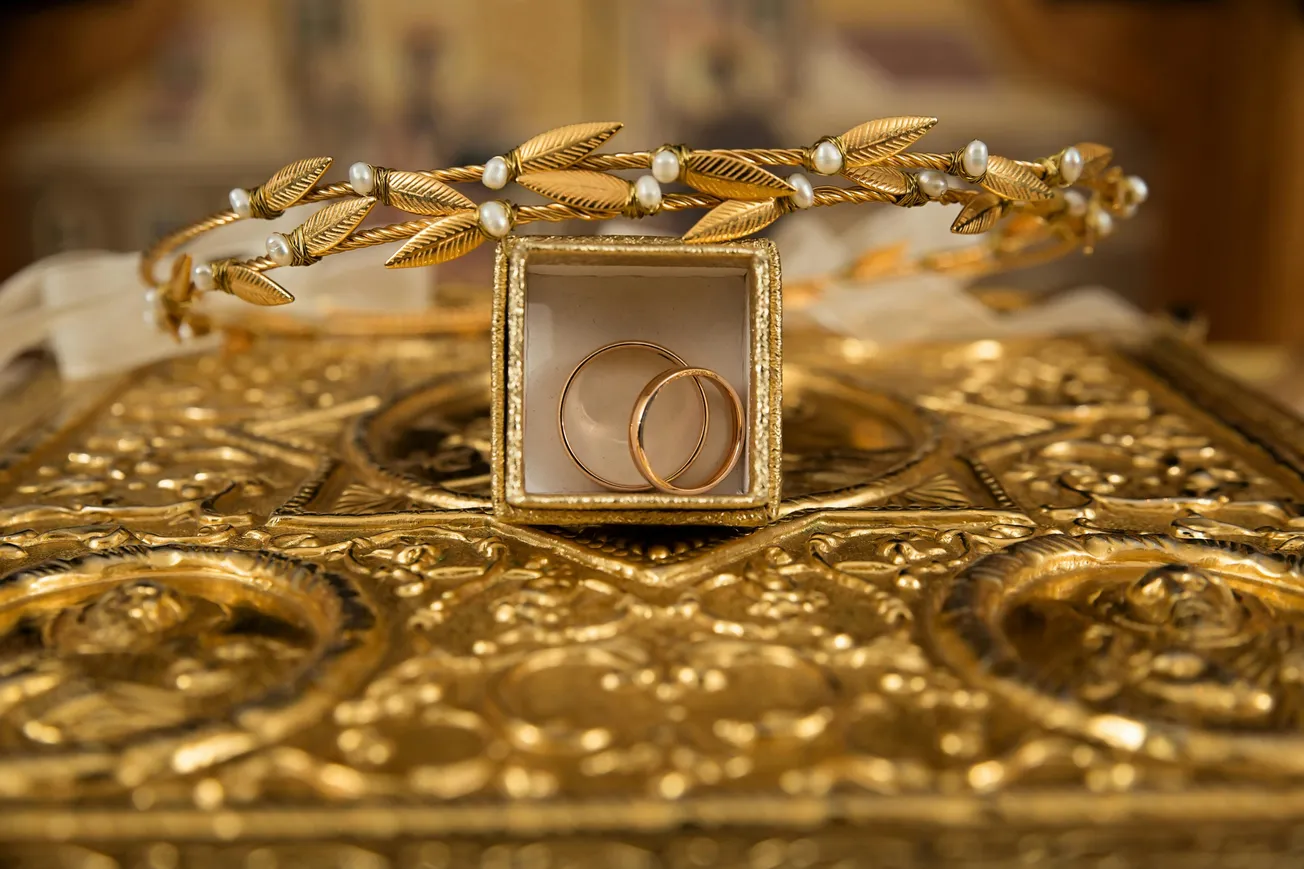 A gold leaf headband adorned with pearls encircles a small box containing two gold rings, resting on an intricately detailed golden surface. Elegant and opulent.