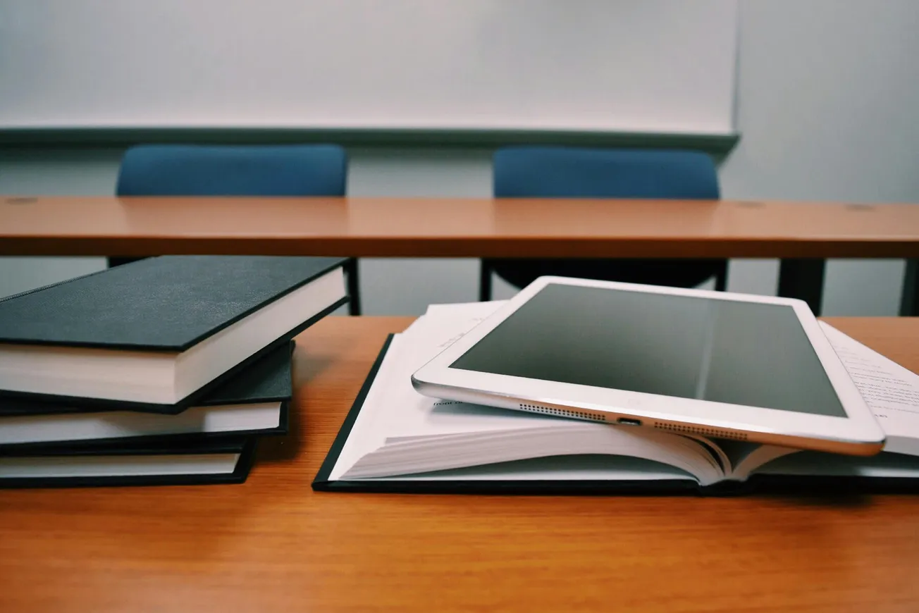 Tablet rests on an open book atop a wooden desk in a classroom. Nearby, stacked closed books. Blue chairs and a whiteboard in the background suggest a study setting.