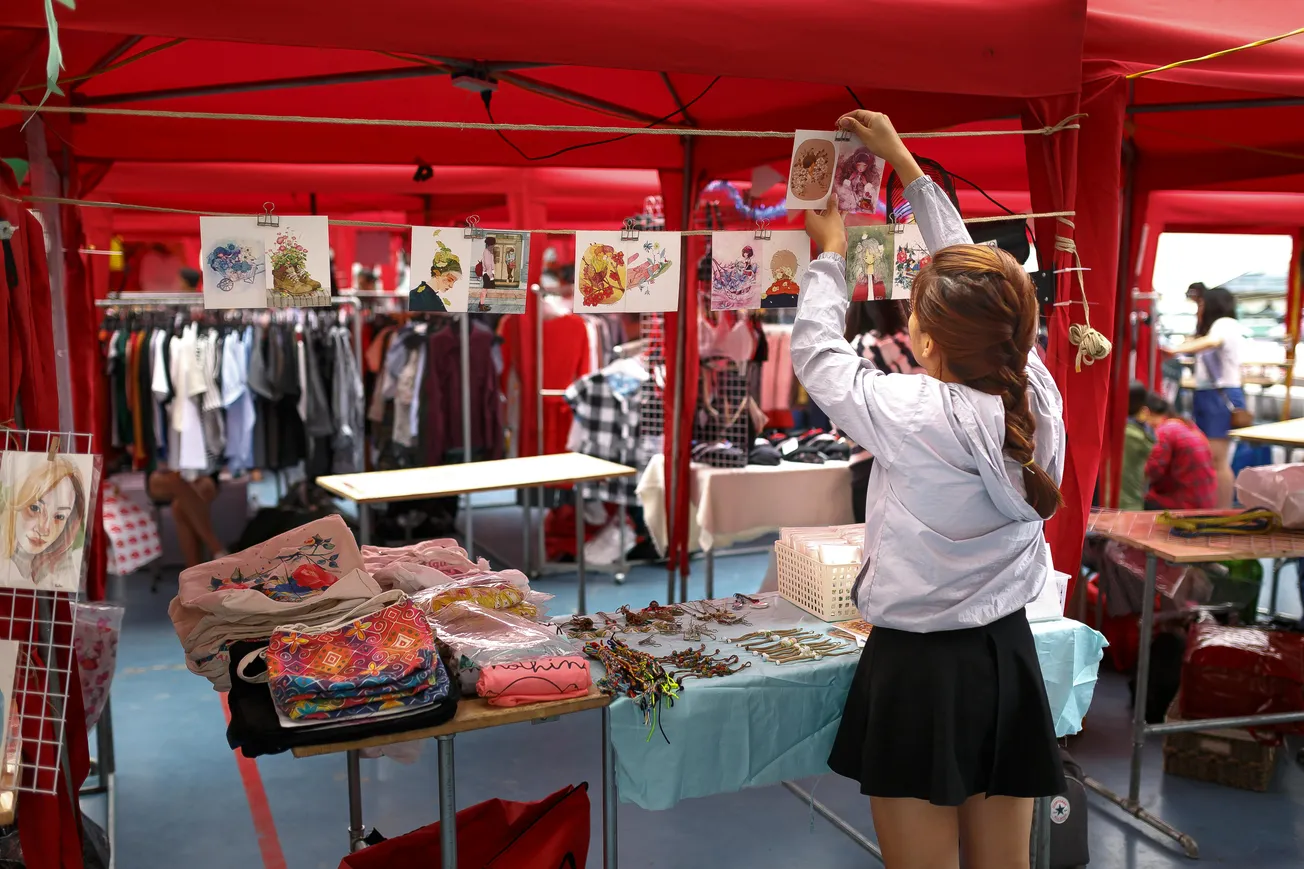 A woman at an outdoor market arranges colorful drawings under a red tent. Clothing racks and folded textiles create an inviting, creative atmosphere.