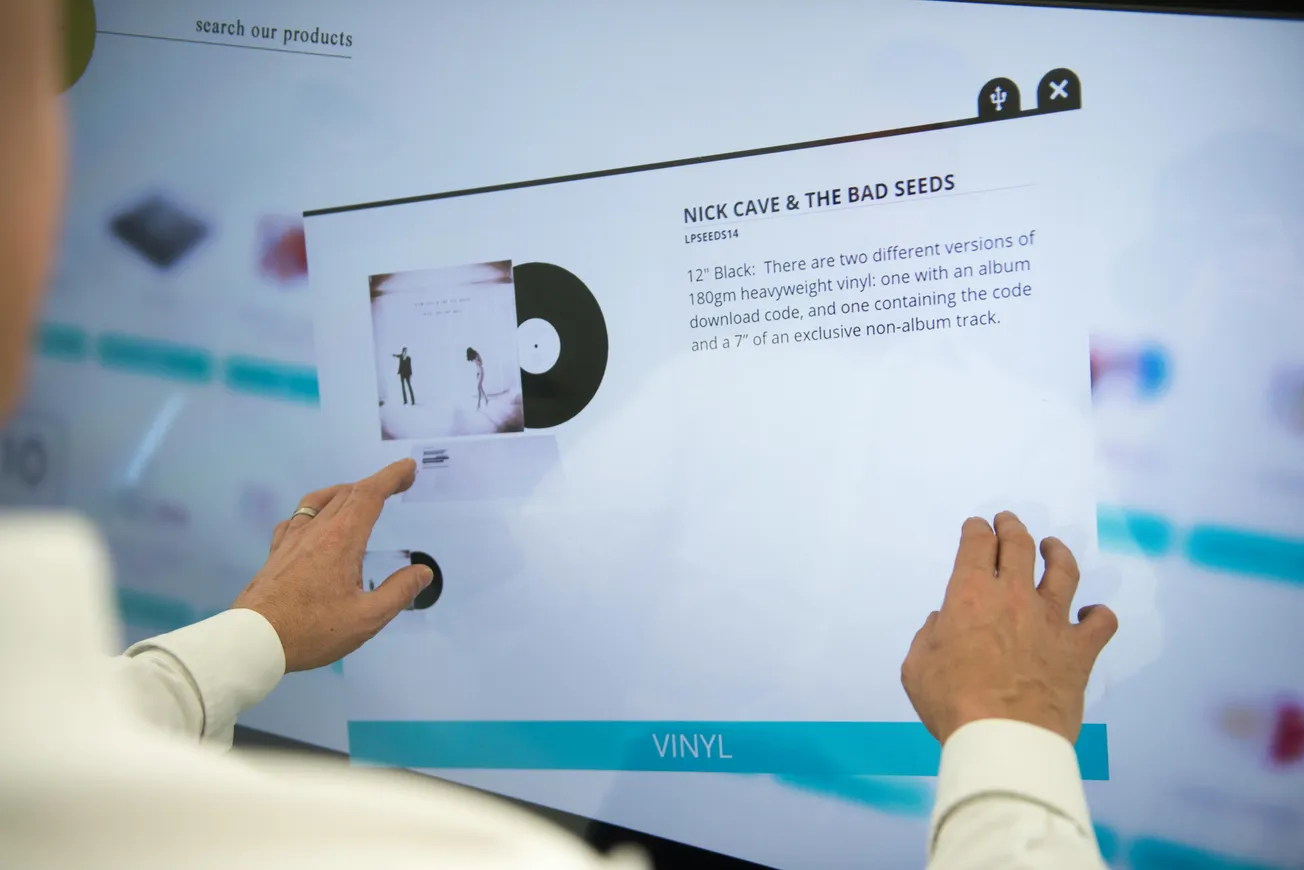 A person interacts with a digital touchscreen displaying information about a Nick Cave & the Bad Seeds vinyl record.