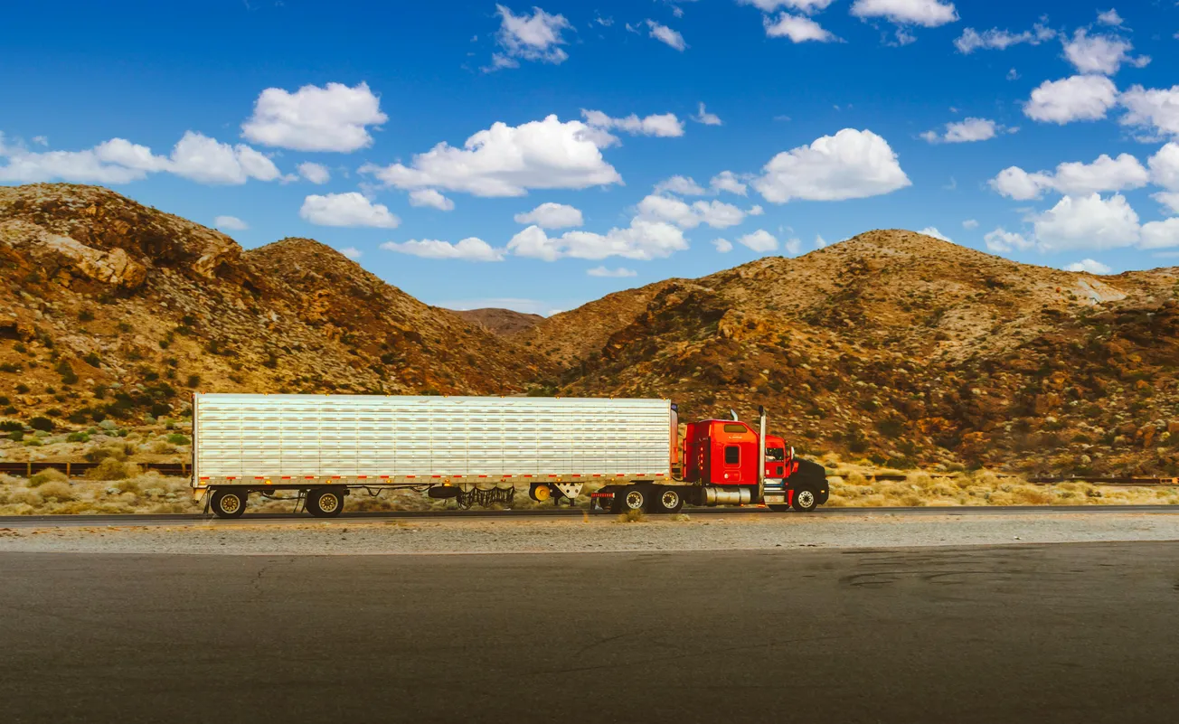 A red semi-truck with a white trailer parked on a highway, set against a backdrop of arid, rocky hills and a blue sky with scattered clouds, conveys a sense of solitude and travel.