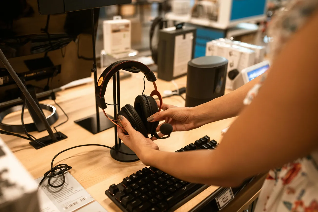 A person examines black and red headphones on a wooden store display next to a monitor and keyboard. The scene suggests a tech shopping experience.