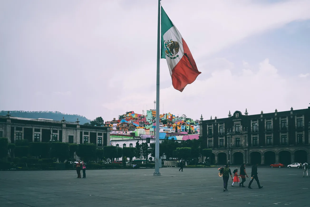 A large Mexican flag waves prominently in a plaza, surrounded by historic buildings. In the background, colorful hillside houses add vibrancy on an overcast day.