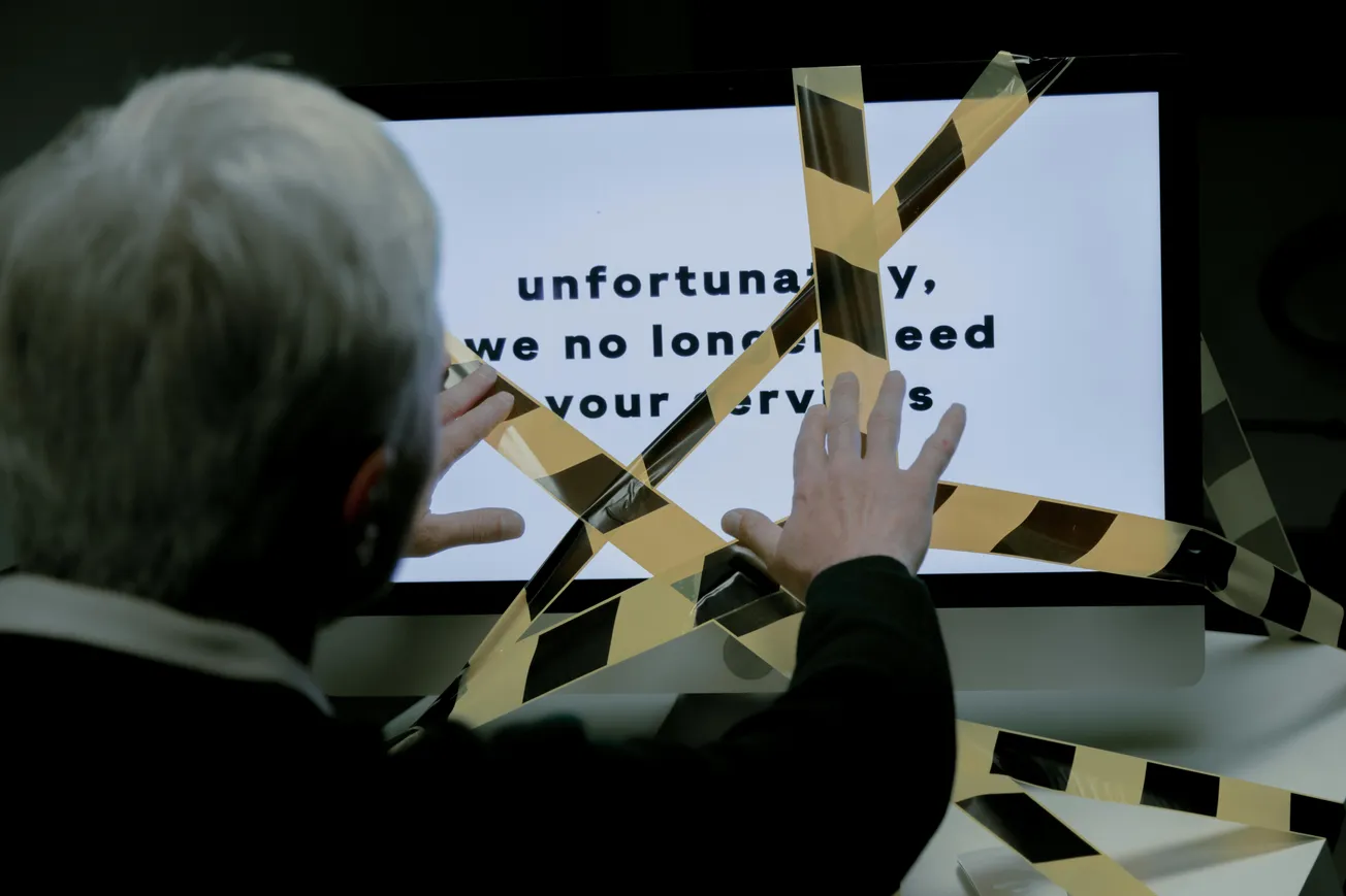A person with gray hair tries to touch a computer screen displaying "unfortunately, we no longer need your services," with caution tape blocking it.