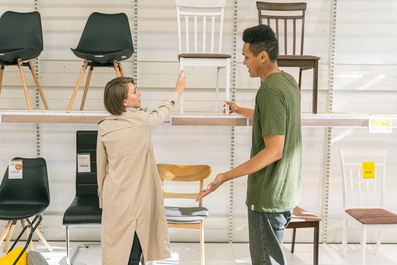 A man and woman in a furniture store discuss chairs on display. The woman in a beige coat points at a white chair. They appear engaged and thoughtful.