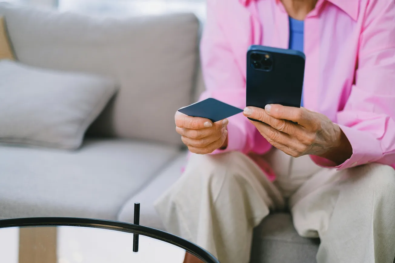 A person in a pink shirt sits on a sofa, holding a smartphone and a credit card, suggesting online shopping. The setting is casual and relaxed.