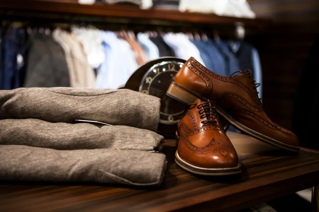 Brown leather dress shoes and folded gray blazers rest on a wooden table. A vintage clock and hanging clothes form the blurred background, conveying elegance.