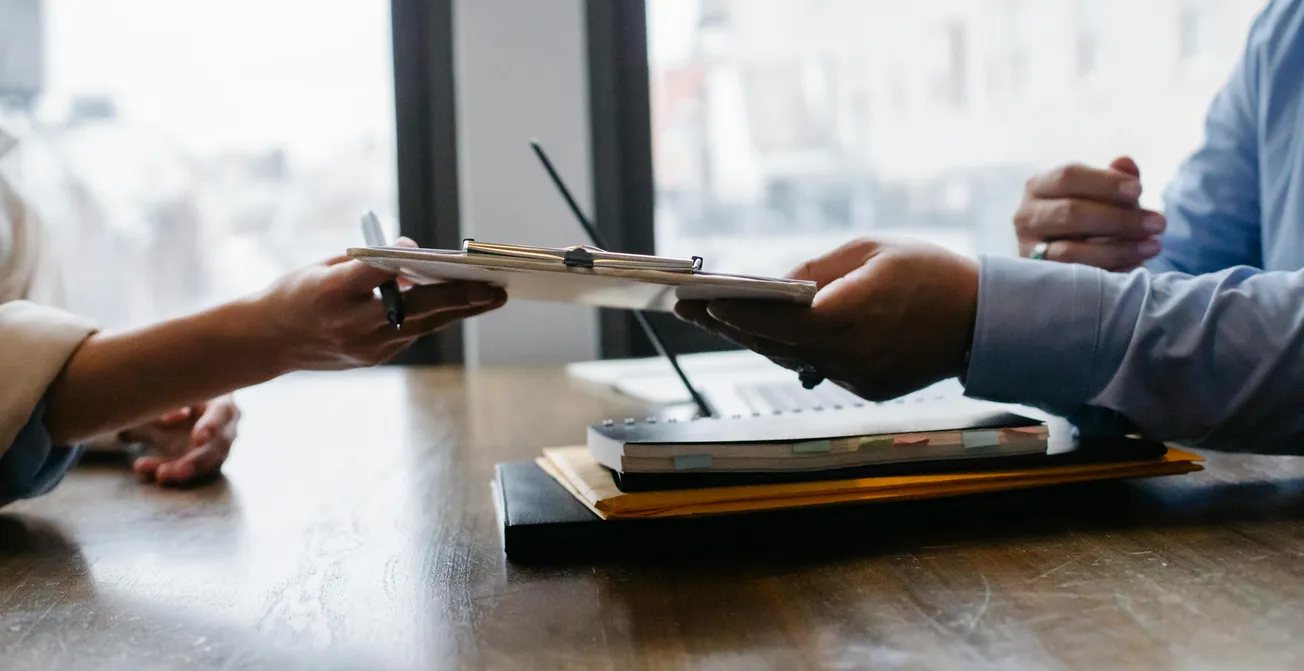 Two people seated at a wooden table exchange documents. One hand holds a pen, suggesting a formal or business context. The mood is professional.