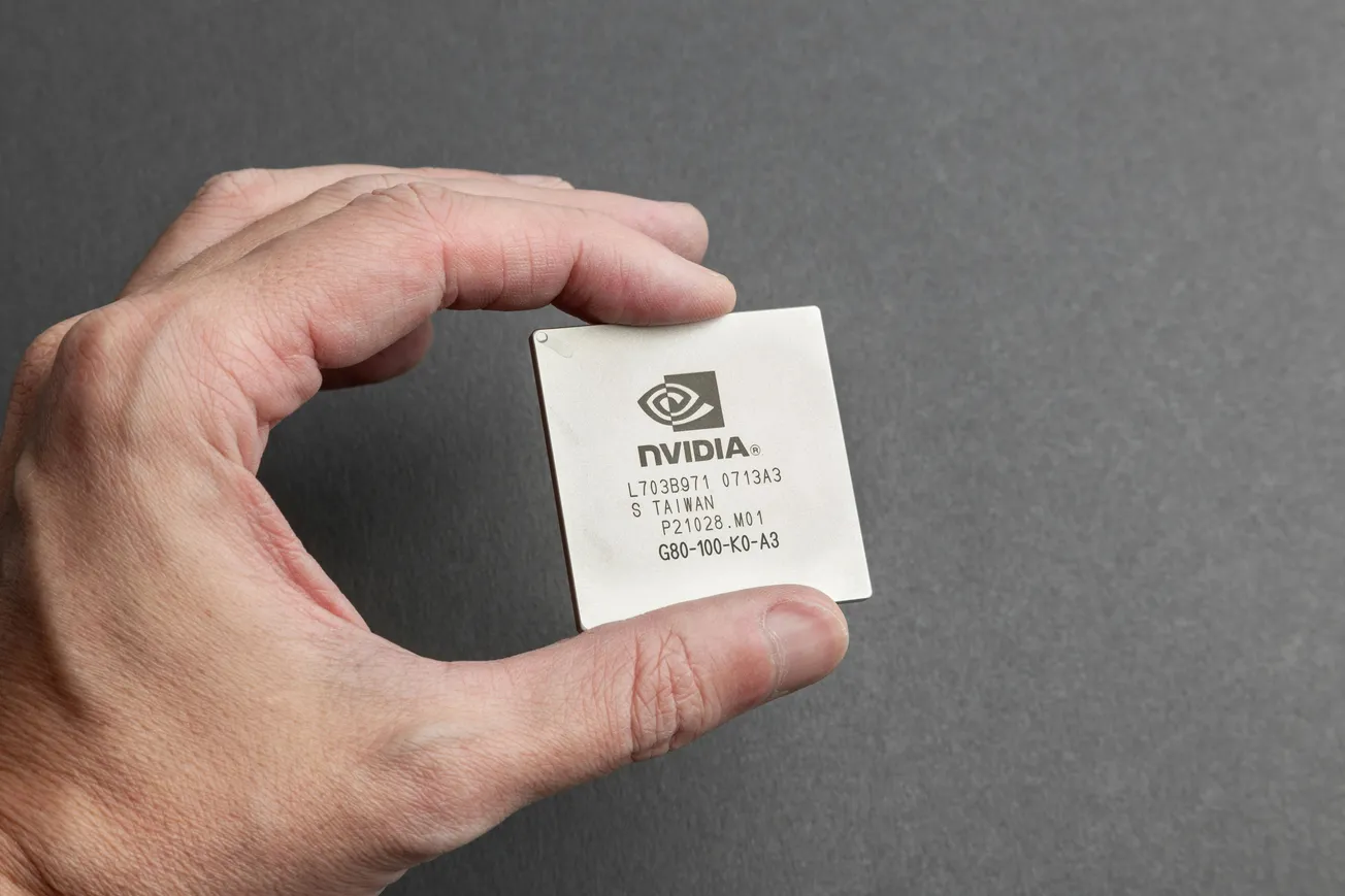 A hand holding an NVIDIA computer chip against a gray background. The chip is square, metallic, and engraved with technical specifications.