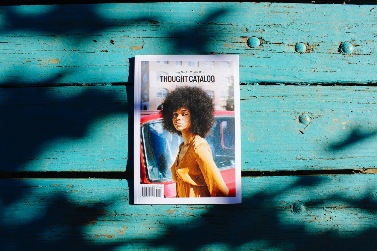 A magazine titled "Thought Catalog" featuring a woman with curly hair in a yellow top is placed on weathered, teal wood, with artistic shadows.