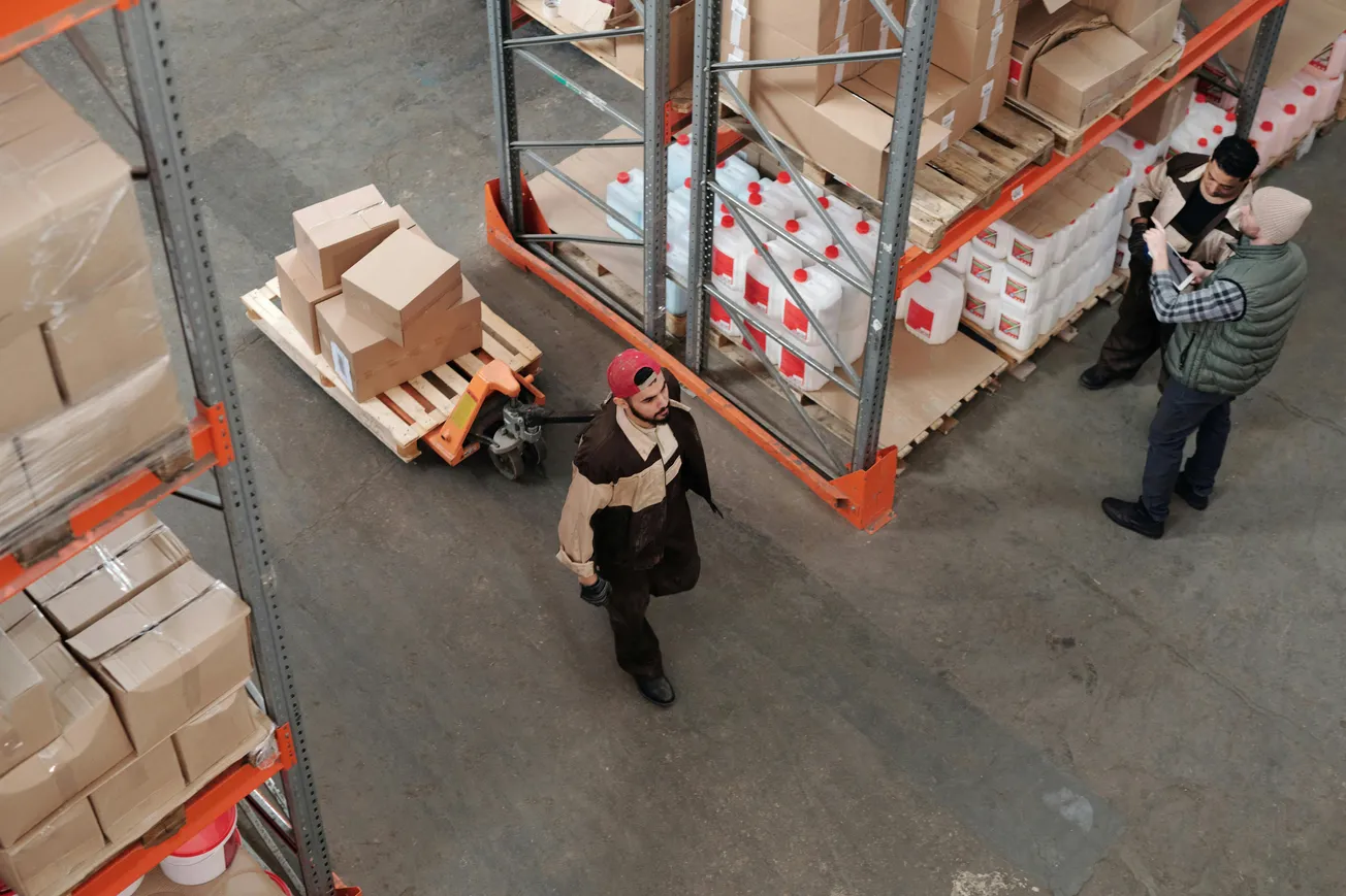 Warehouse scene from above showing a worker in a red cap pulling a pallet jack with boxes. Two others stand nearby, reviewing a clipboard.