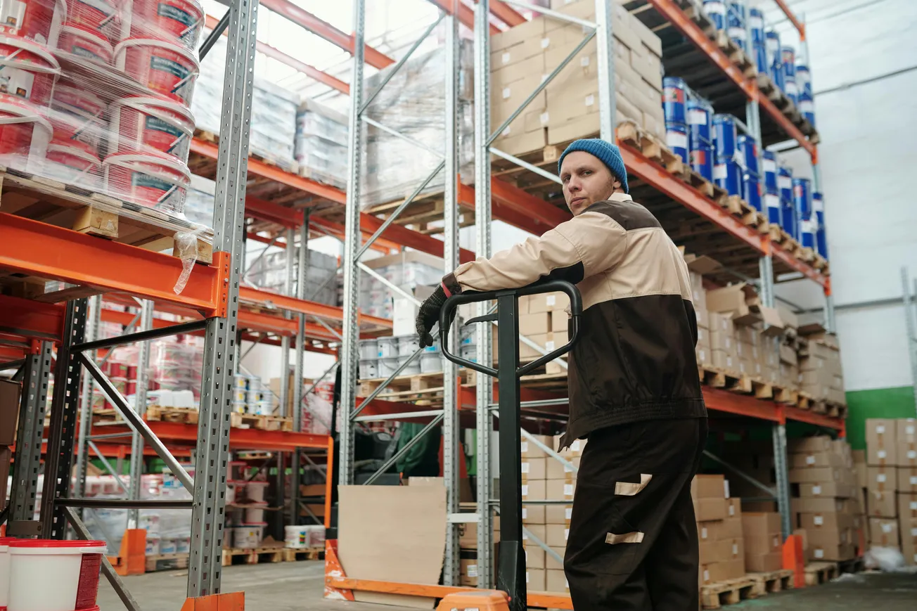 A warehouse worker in a brown uniform and blue beanie stands with a pallet jack. He is surrounded by shelves of boxes and paint buckets. The setting feels industrious and organized.