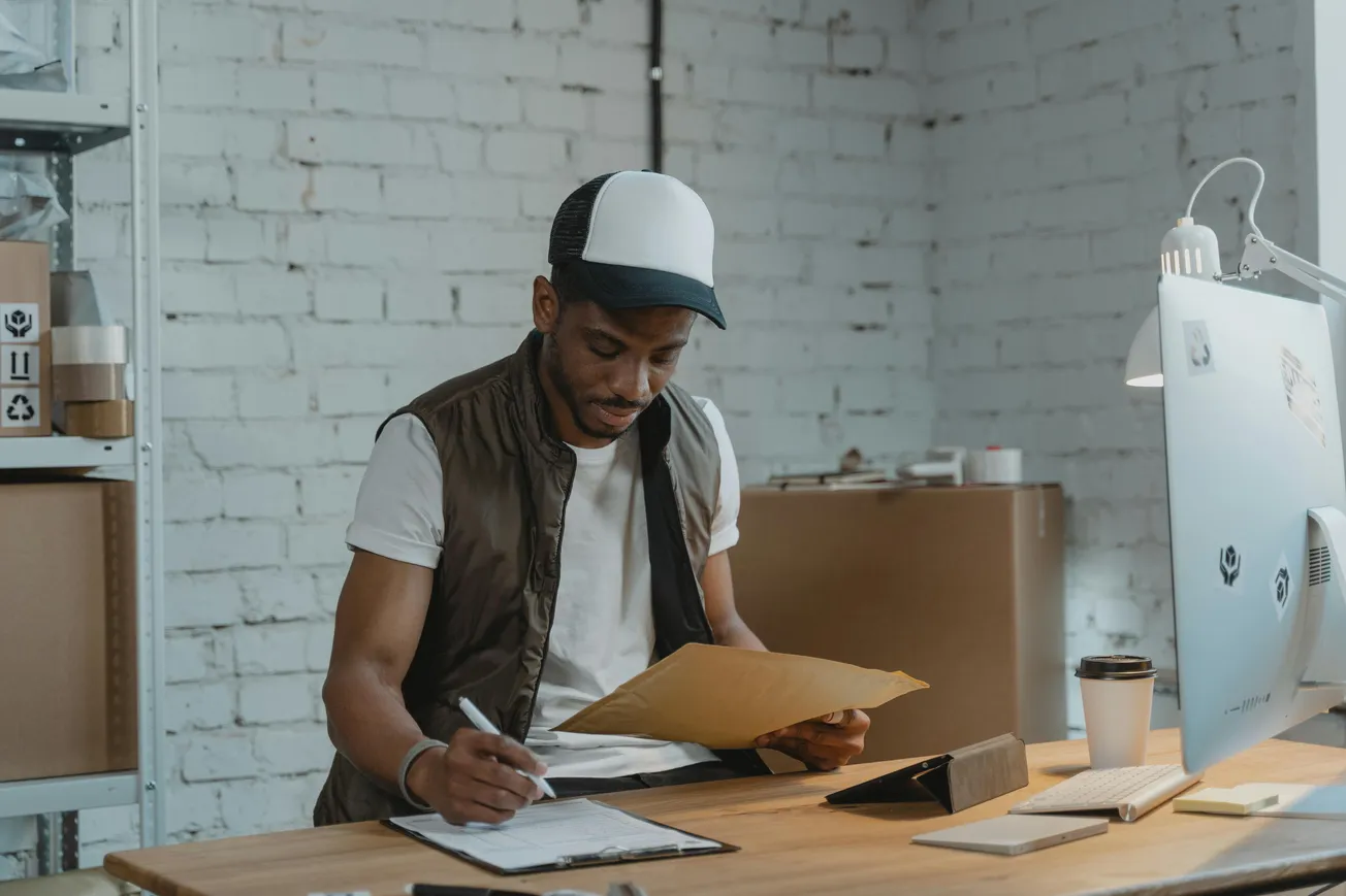 A man in casual attire and a cap sits at a desk, focused on paperwork in a bright office with a modern computer and coffee cup nearby.