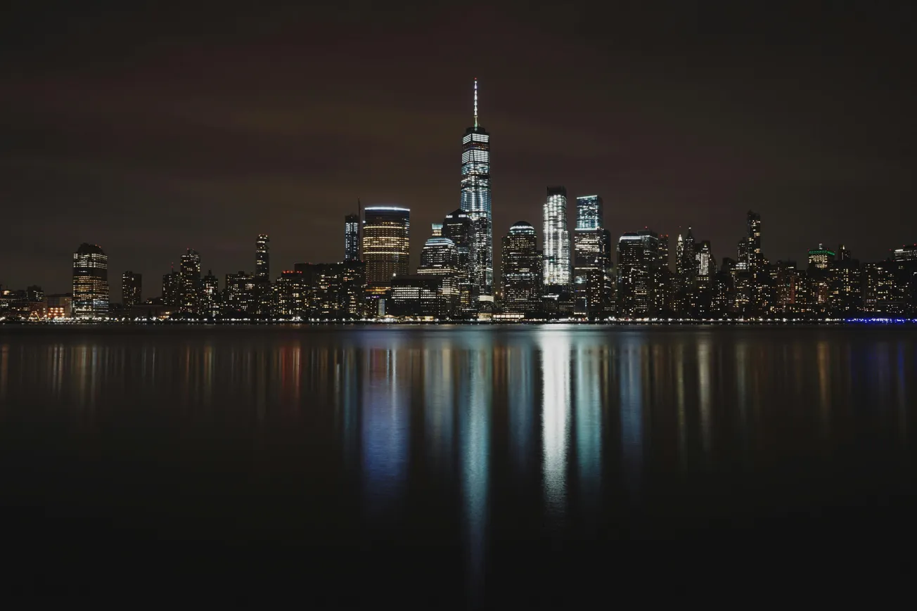 Nighttime city skyline with illuminated skyscrapers reflecting on calm water. The tallest building stands prominently in the center, set against a dark sky.